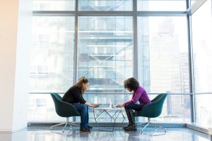 two colleagues in a workplace, leaning over a table having a conversation.